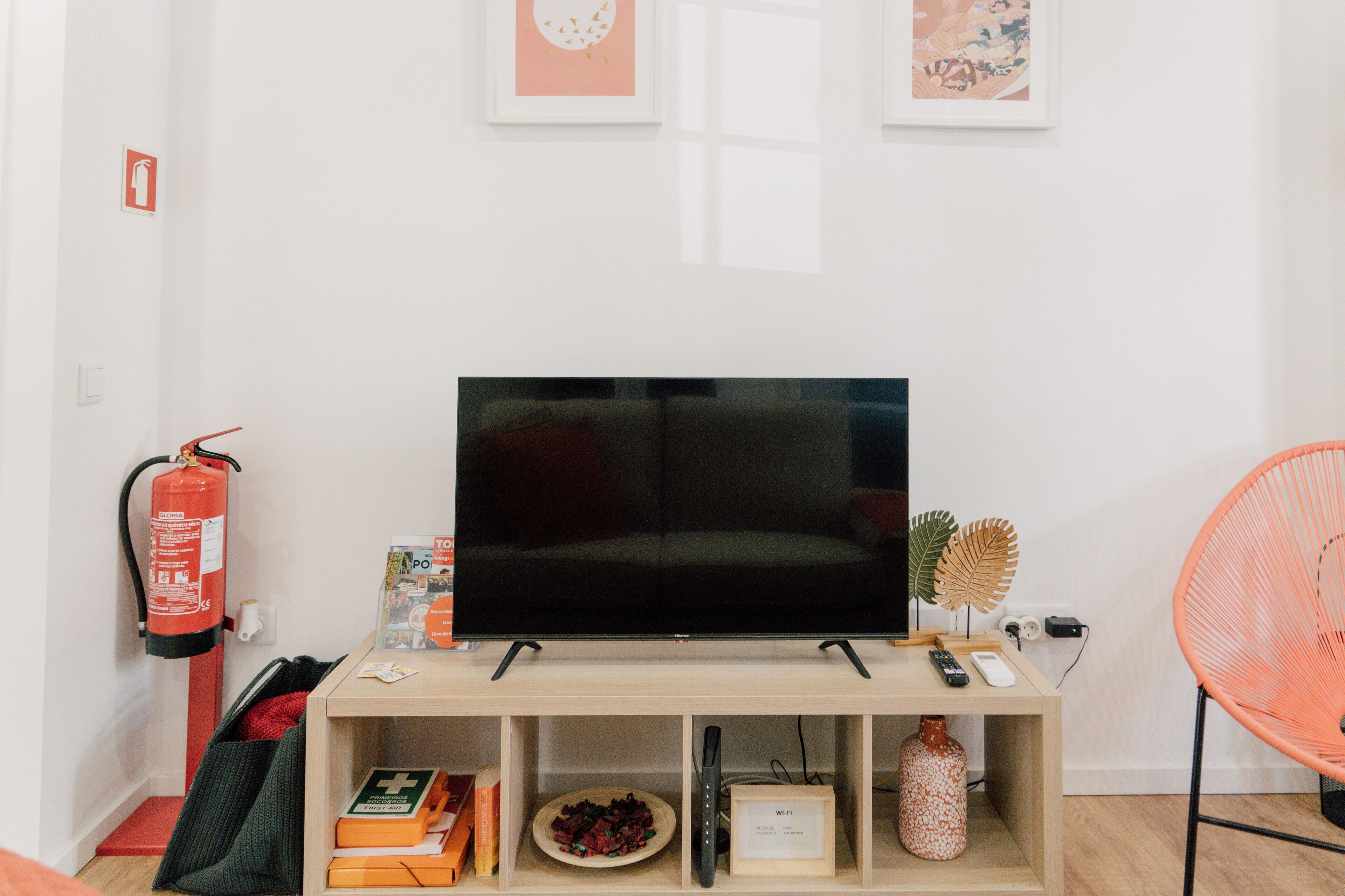 A living room with a flat-screen TV on a wooden stand, a fire extinguisher, a bookshelf with books and decorative items, and a red chair.