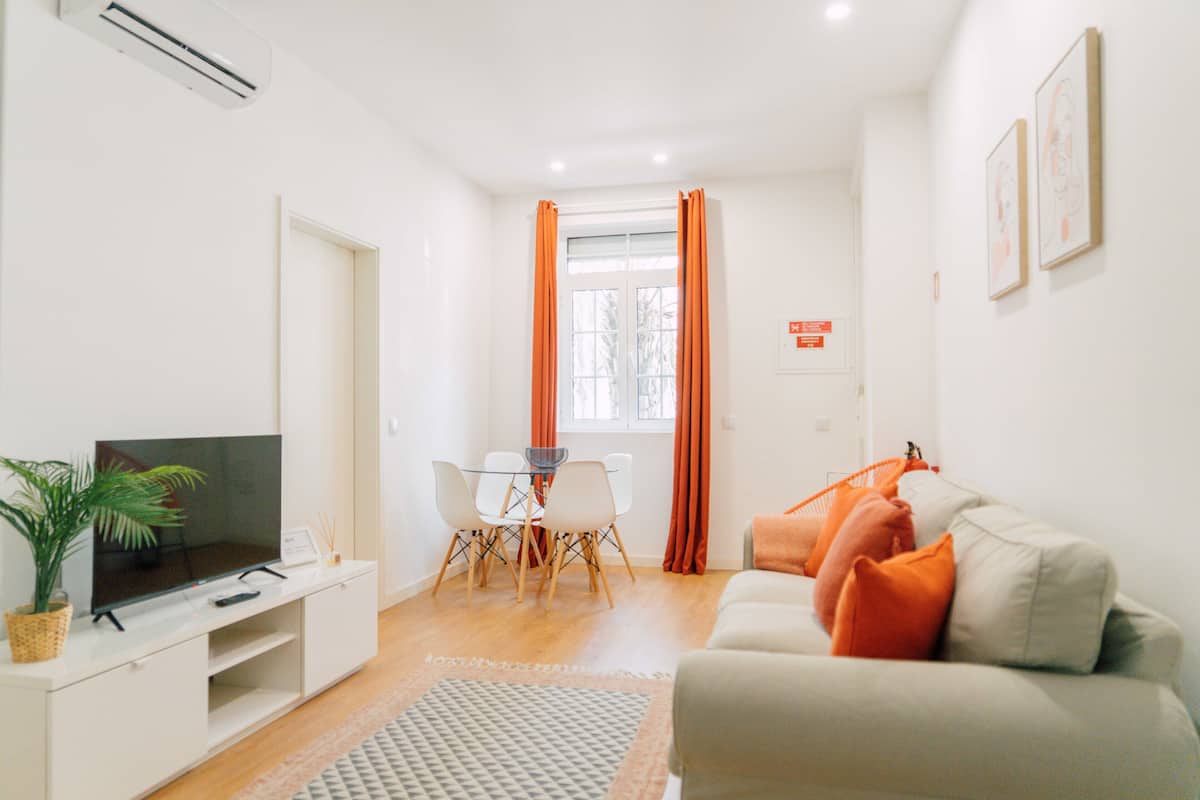 A modern living room with a flat-screen TV, a dining table with chairs, a sofa with orange cushions, and a window with orange curtains.