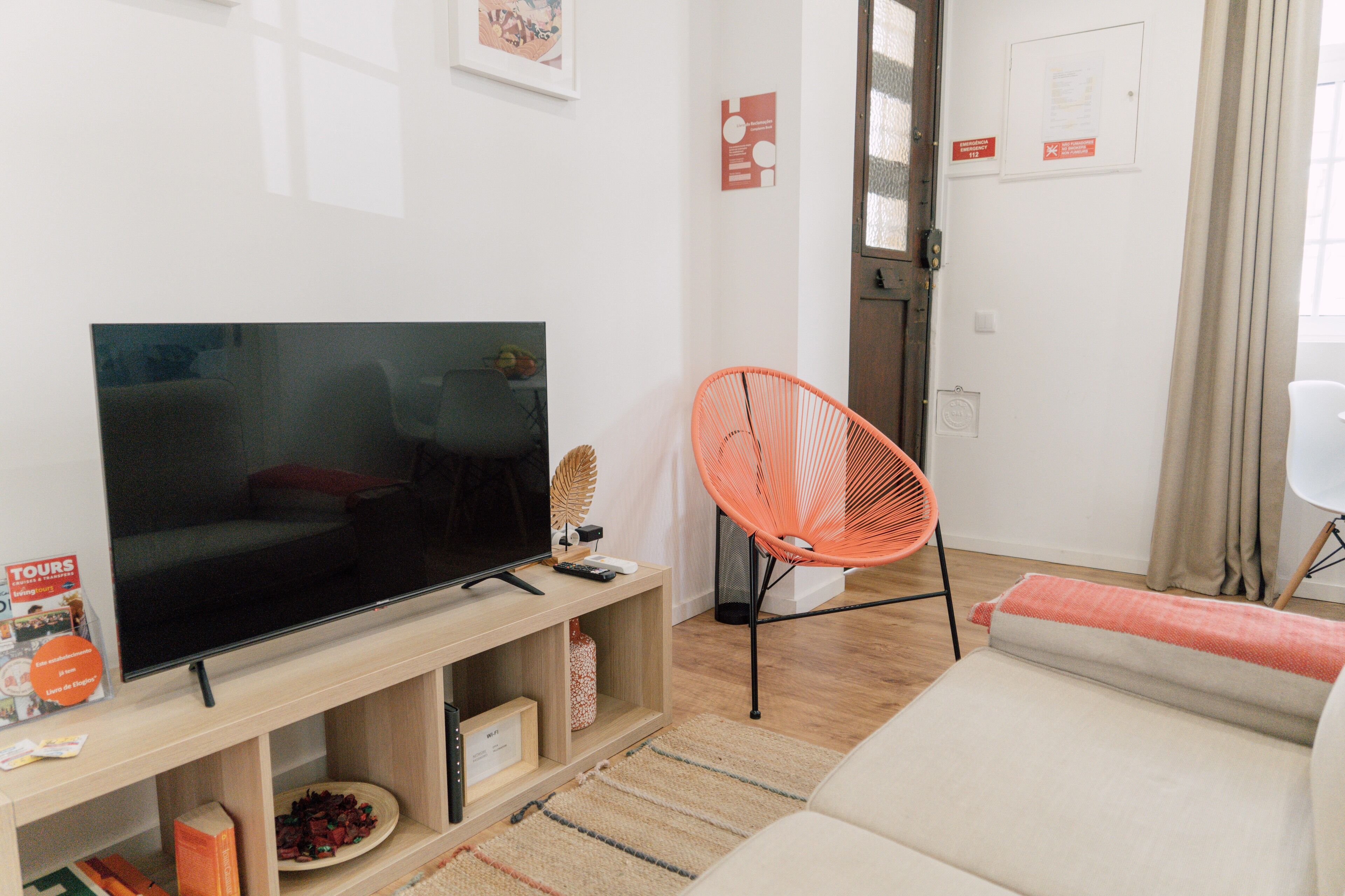 A living room with a flat-screen TV on a wooden stand, a bright orange chair, a sofa, and a small table with a bowl of fruit.