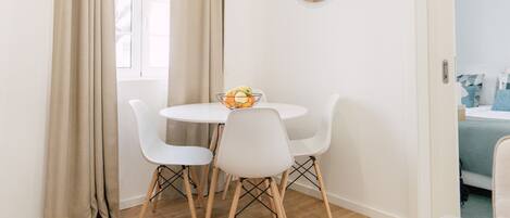 A dining area with a round table, four white chairs, a bowl of fruit, and a wall clock.