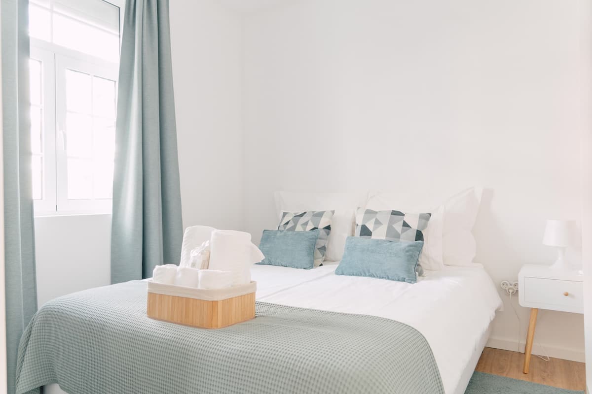 A neatly arranged bedroom with a bed, pillows, a bedside table, and a window with curtains.