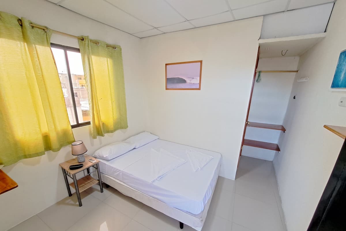 Een slaapkamer met een bed, een nachtkastje, een lamp, een raam met gordijnen, een lijst met een afbeelding en een trap.