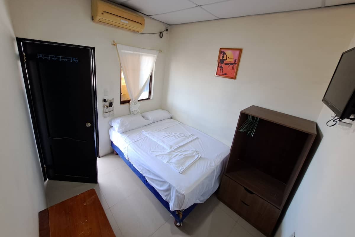 Een kleine, schone kamer met een eenpersoonsbed, een houten garderobe, een tv aan de muur en een plafondventilator.