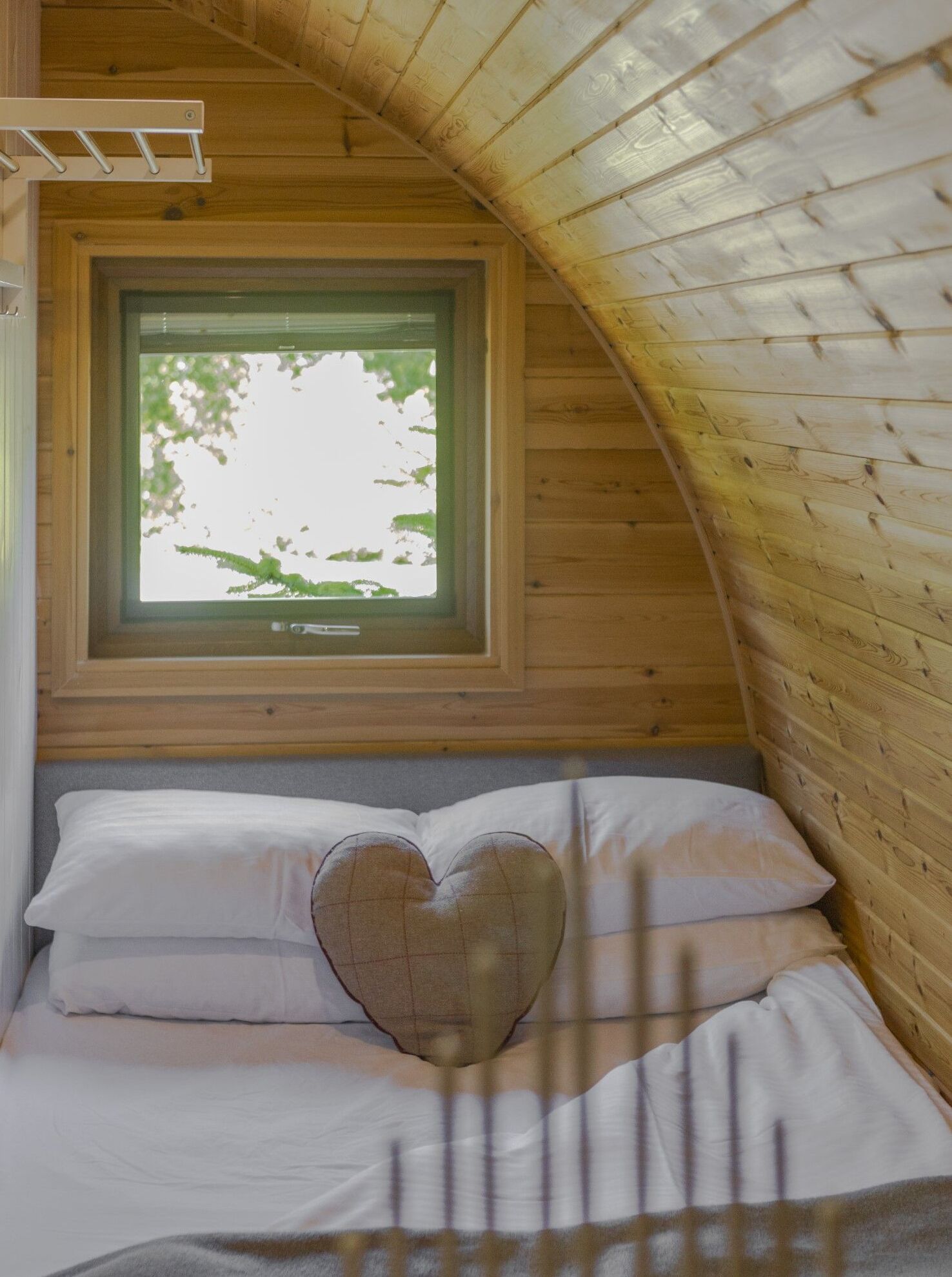 A cozy bedroom with a sloped ceiling, a window with a view of trees, a heart-shaped pillow, and a bed with white linens.