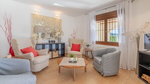 A modern living room with a sofa, armchairs, a coffee table, and a TV. There is a painting on the wall, a window with curtains, and a view of greenery outside. - Villa Isabel by Sun & Sea Homes (Moraira)