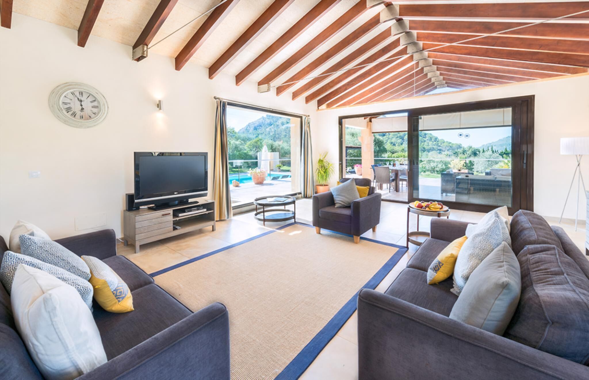 A modern living room with a flat-screen TV, two sofas with cushions, a coffee table, and a large window offering a view of a pool and mountains. - Villa S'airieta - Finca-Style Villa with Private Gardens Near Playa de Muro (Mallorca)