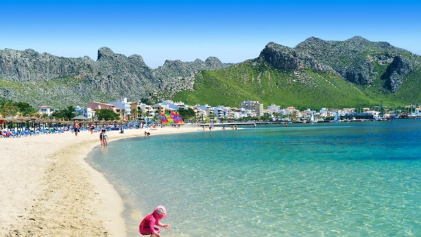 A beach with clear turquoise water, a sandy shore, and a backdrop of rocky mountains. - Villa S'airieta - Finca-Style Villa with Private Gardens Near Playa de Muro (Mallorca)