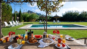 A breakfast table with croissants, bread, fruit, and beverages, overlooking a swimming pool and lounge chairs.