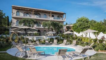 Un espace piscine doté de chaises longues et de parasols, entouré d’une végétation luxuriante et bordé d’un bâtiment comportant de nombreux balcons.