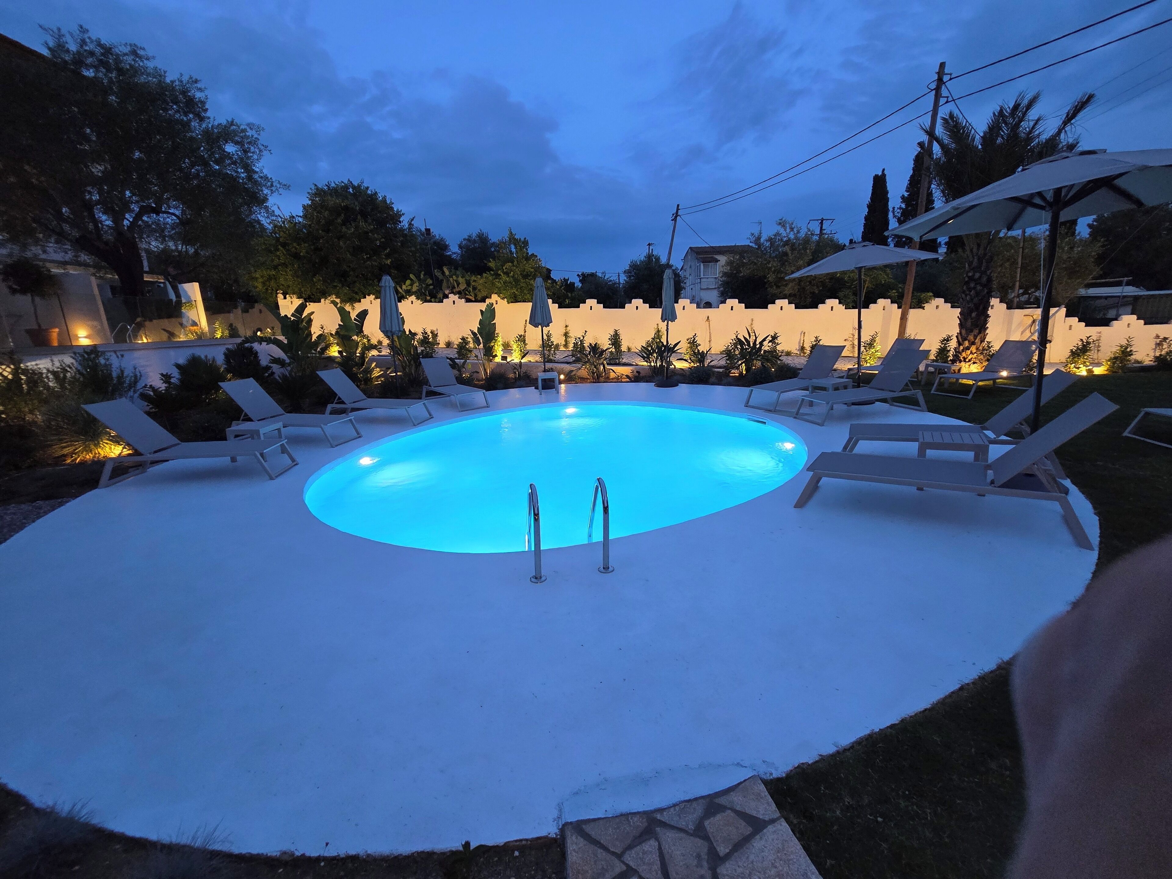 A circular pool with a blue-lit edge, surrounded by white lounge chairs and umbrellas, set against a backdrop of trees and a building.
