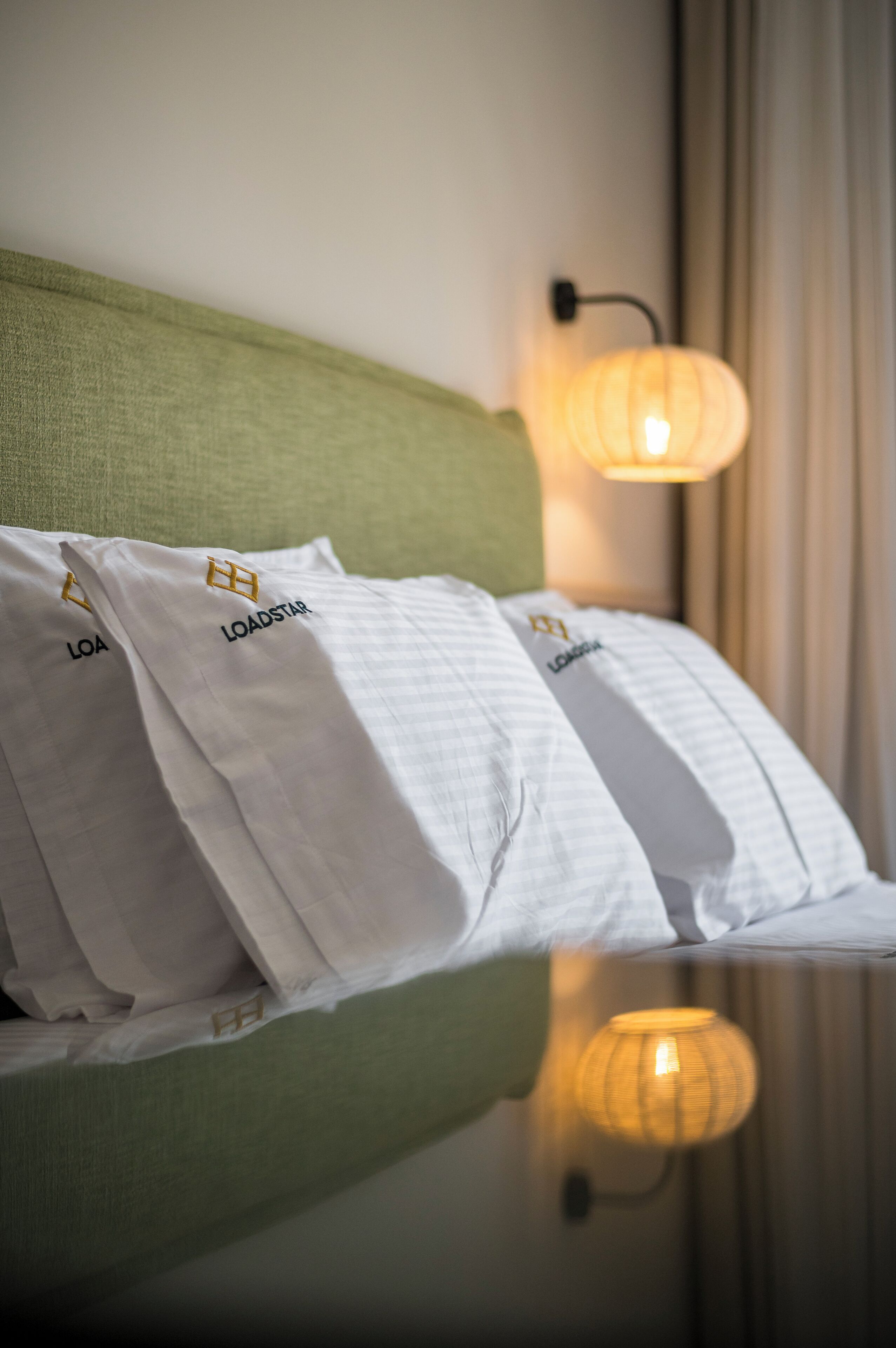 Un lit bien fait, avec des oreillers blancs portant les inscriptions « LOA » et « LOADSTAR », une tête de lit verte et une lampe fixée au mur.