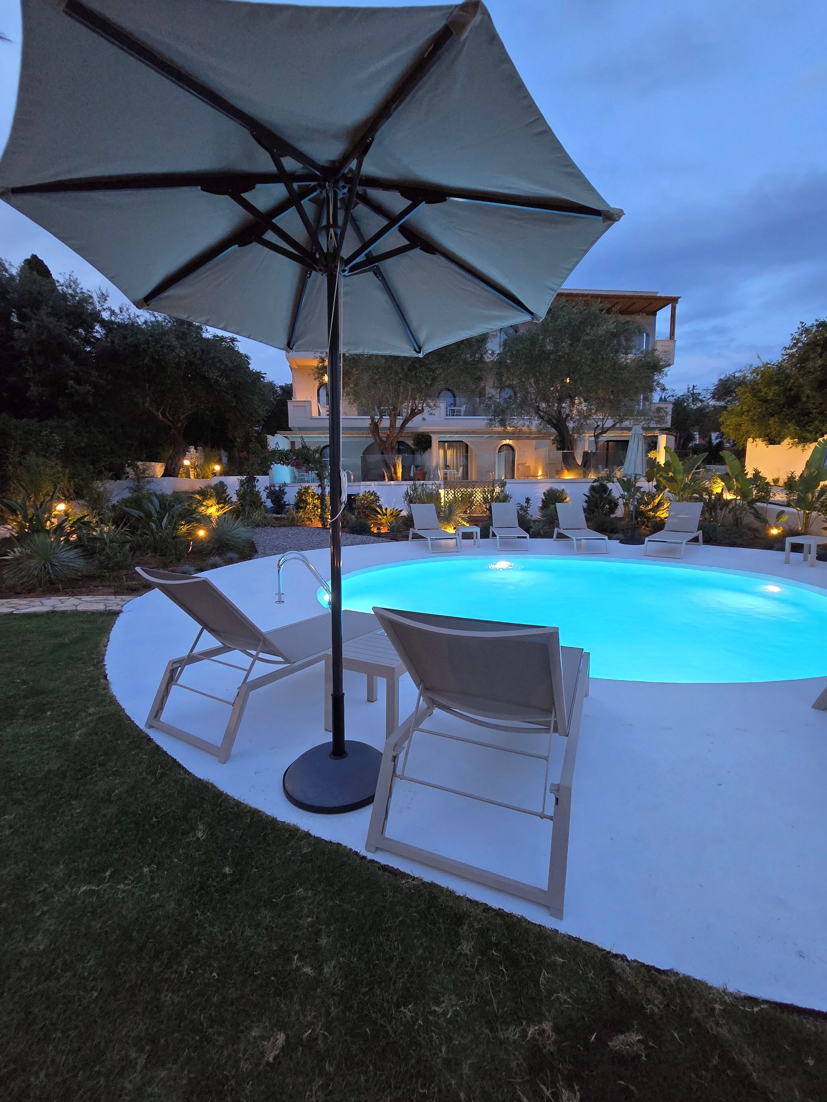 A swimming pool with lounge chairs and an umbrella, surrounded by landscaped garden and a building in the background.