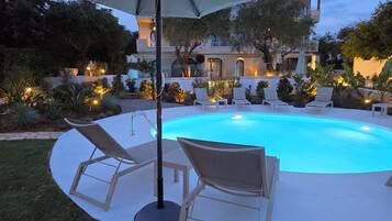A swimming pool with lounge chairs and an umbrella, surrounded by landscaped garden and a building in the background.