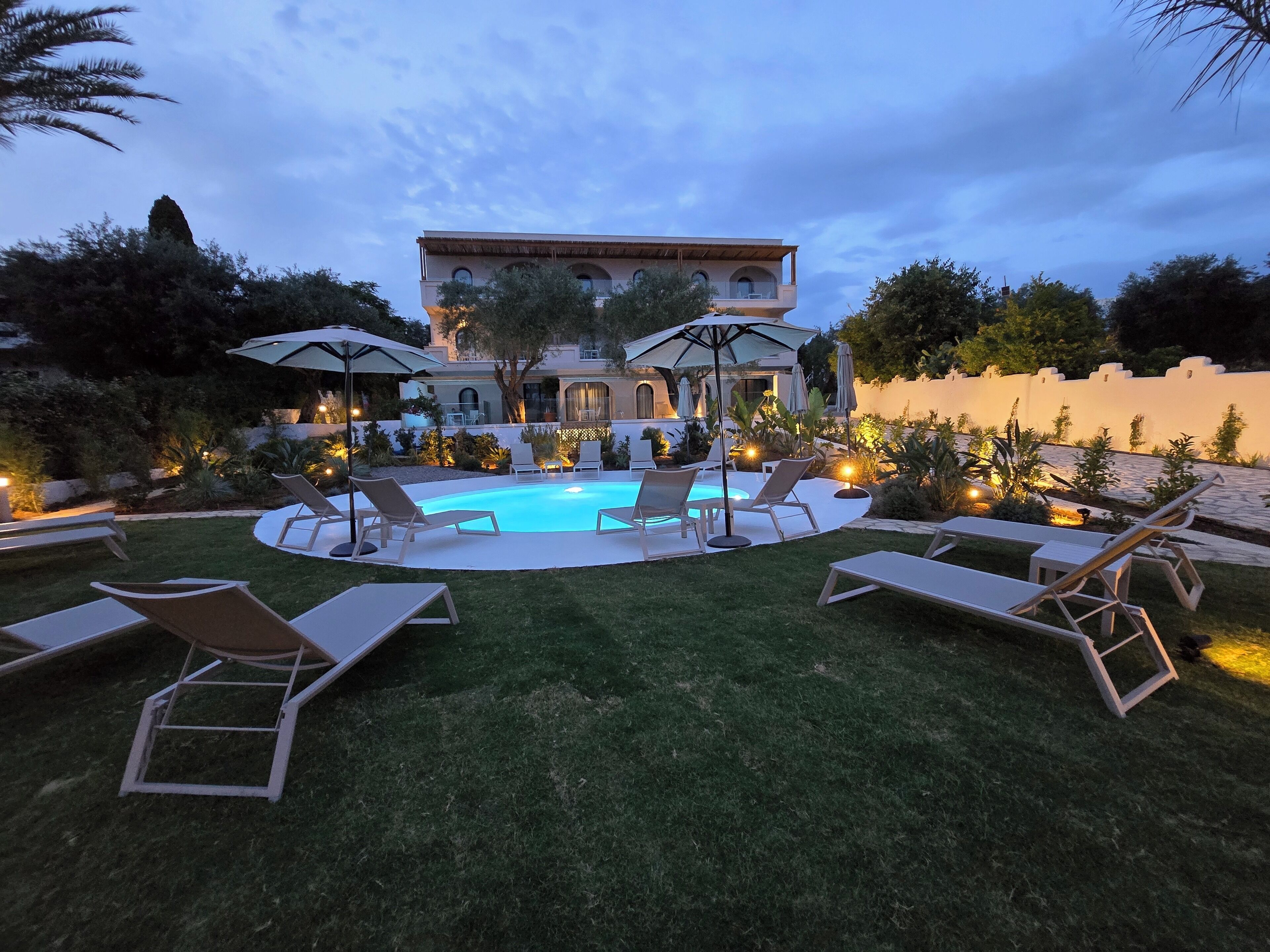 A pool area with lounge chairs and umbrellas, surrounded by a well-maintained lawn and a building with arched windows.