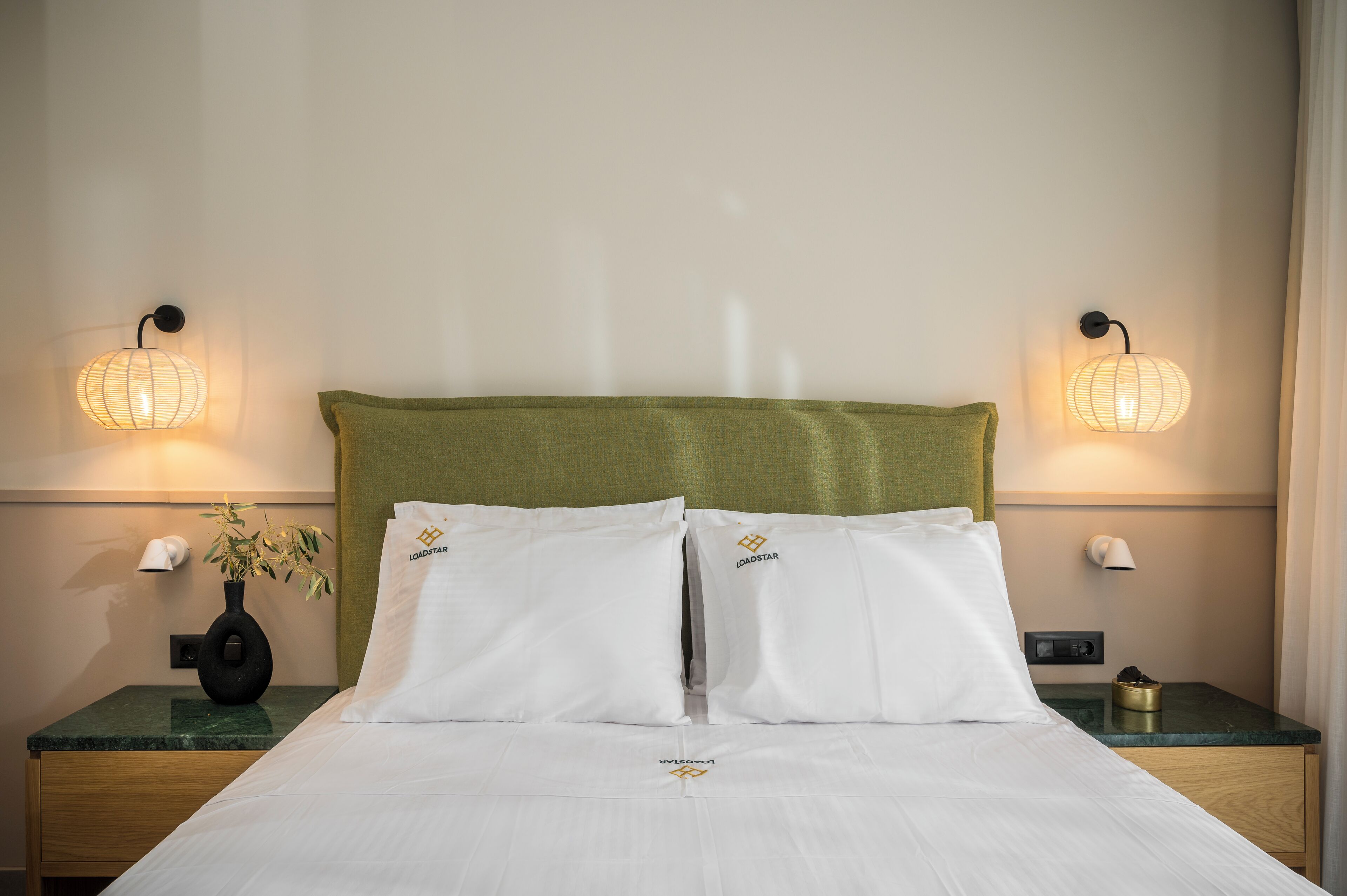 A neatly made bed with white linens, a green headboard, and two pillows with a logo. There are two bedside tables, each with a lamp and a small vase with a plant.