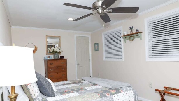 Un dormitorio con cama, cómoda, ventilador de techo, persianas y un pequeño estante de madera.