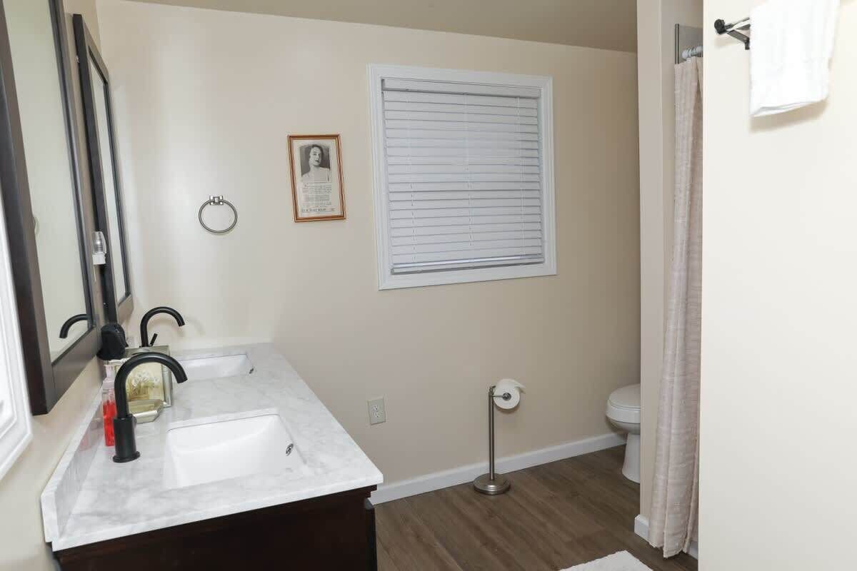 Een badkamer met een witte wastafel, een spiegel, een ingelijste foto, een raam met zonwering, een toilet en een vloerlamp.