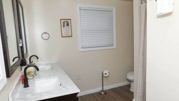 Een badkamer met een witte wastafel, een spiegel, een ingelijste foto, een raam met zonwering, een toilet en een vloerlamp.