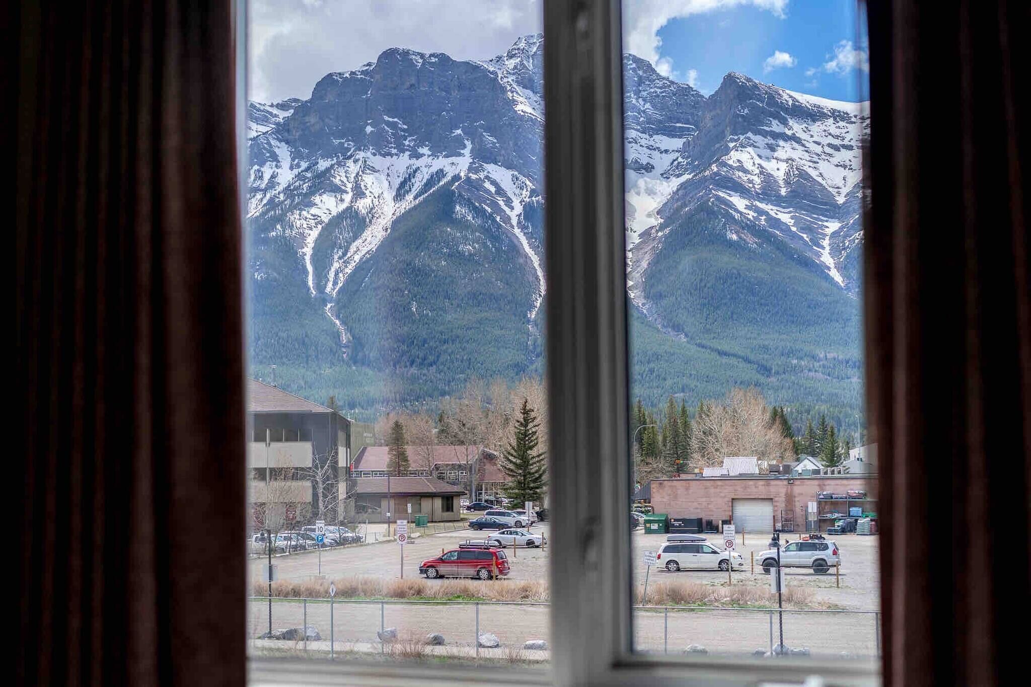 A view through a window of a parking lot with cars, buildings, and snow-capped mountains in the background.