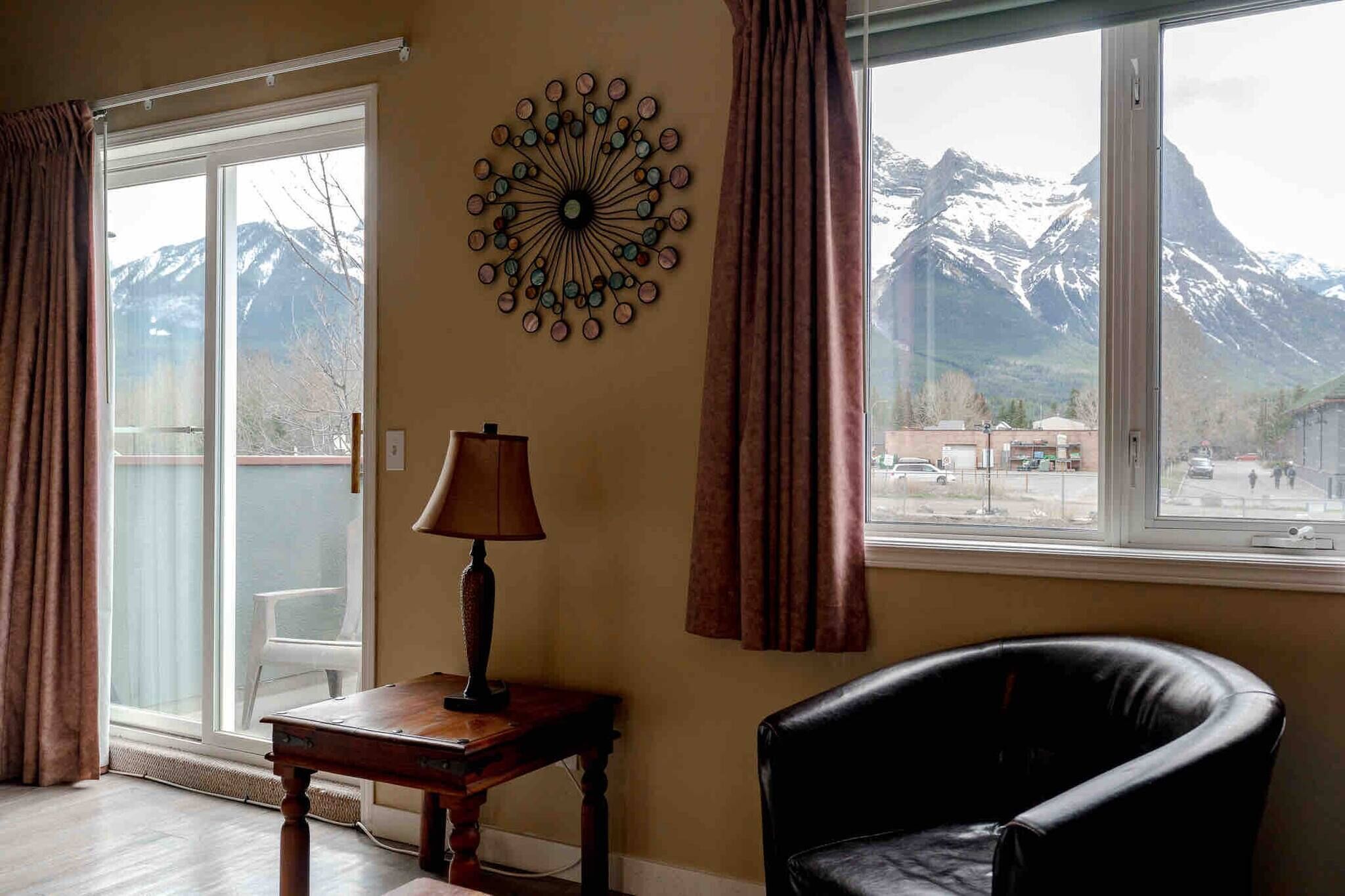 A room with a view of mountains, a lamp, a chair, and a table.