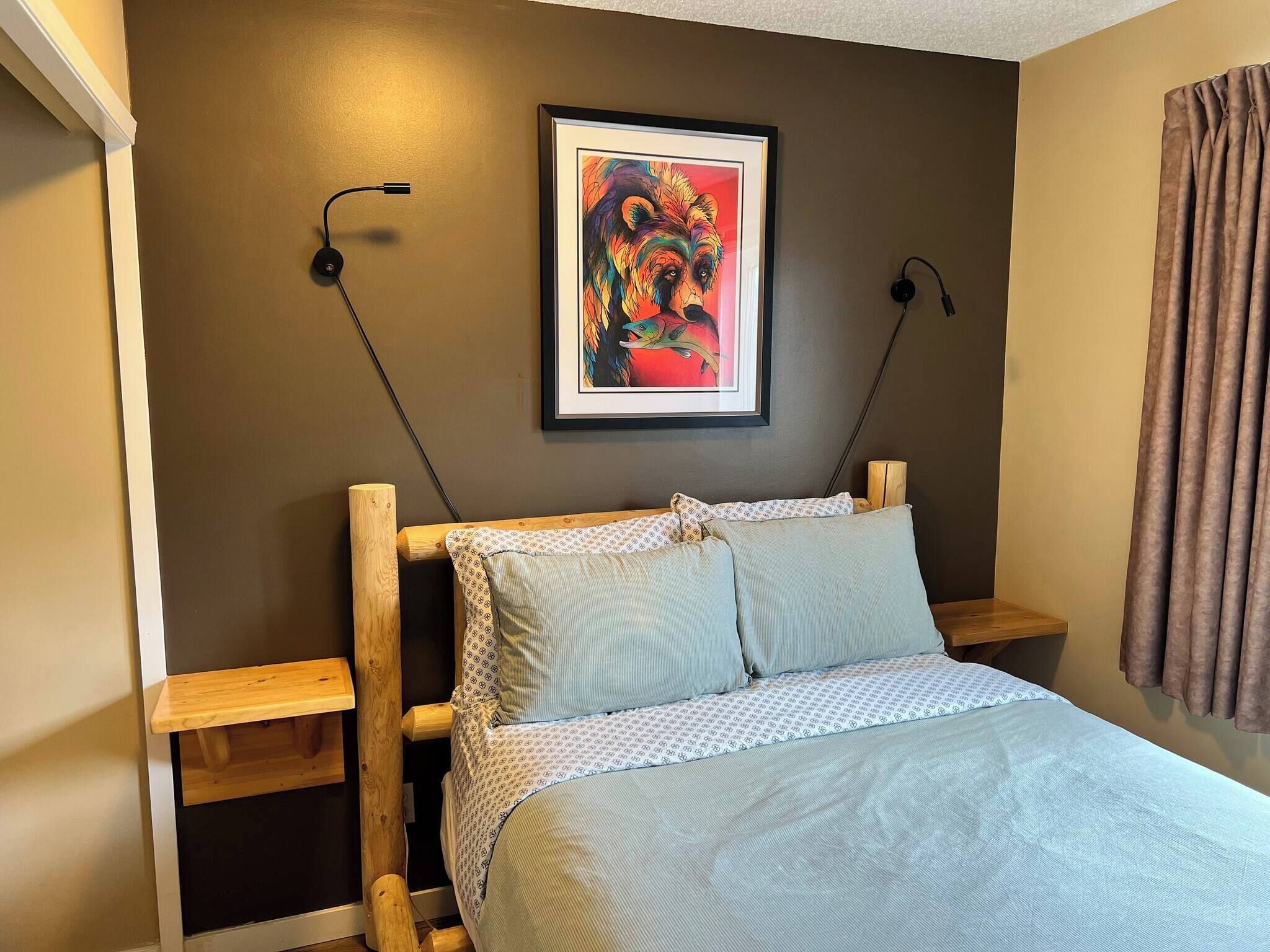 Une chambre à coucher avec une tête de lit en bois, un lit recouvert d’une literie à motifs, un tableau encadré accroché au mur et une table de chevet.
