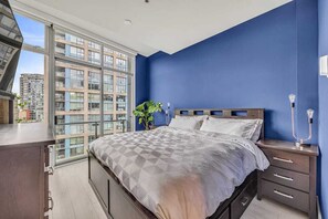 Un dormitorio con una cama grande, una mesita de noche, una lámpara y vistas a edificios a través de un ventanal.