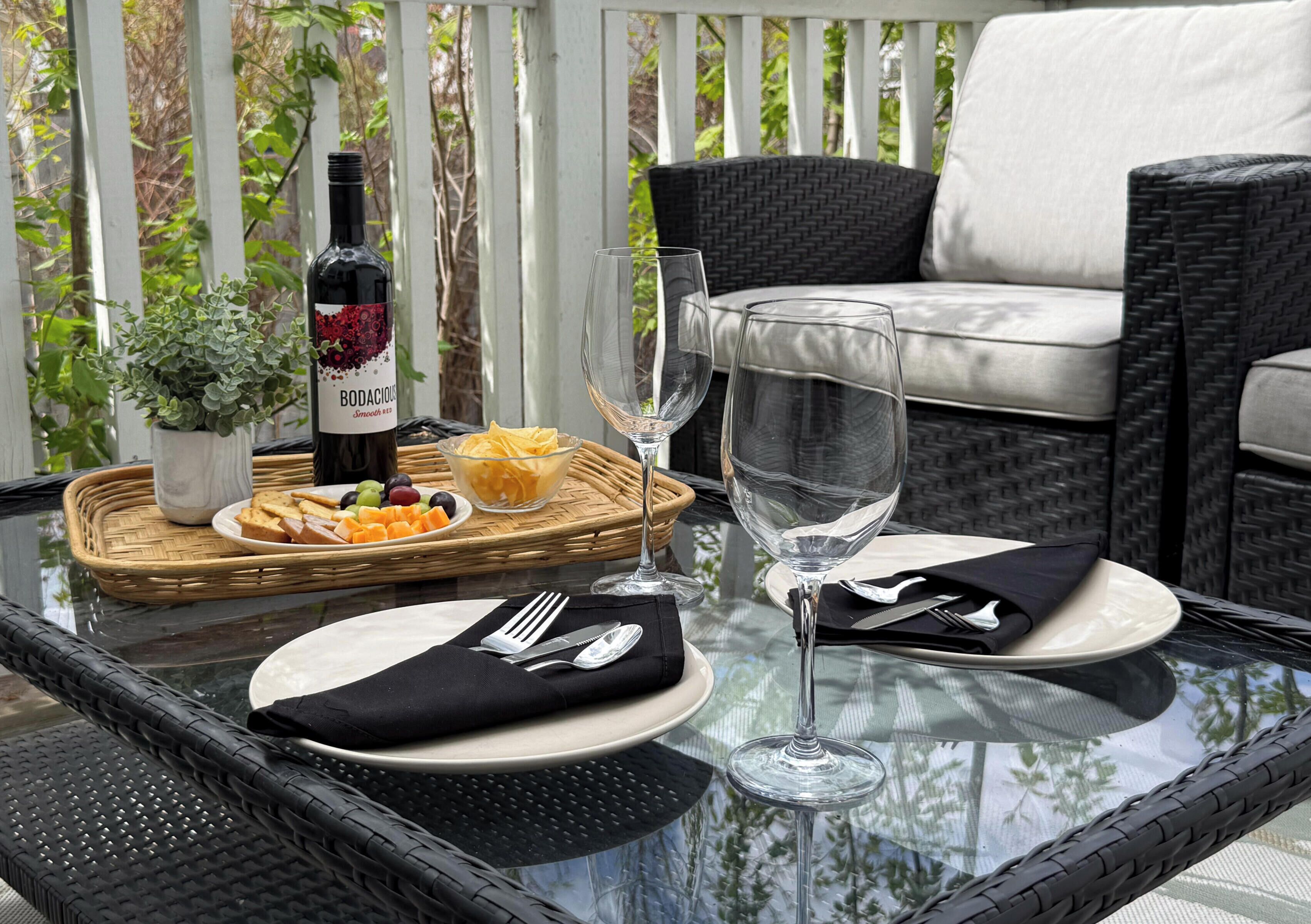 A glass-top table with a bottle of wine, empty wine glasses, plates, cutlery, and a basket of fruit on a balcony.