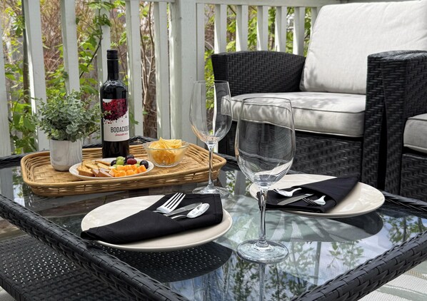 A glass-top table with a bottle of wine, empty wine glasses, plates, cutlery, and a basket of fruit on a balcony.
