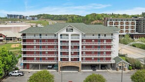 A multi-story hotel with a central entrance, parking lot, and a view of surrounding buildings and greenery. - Wake Up to the River, Walk to the Fun - 3 BR Condo (Pigeon Forge)