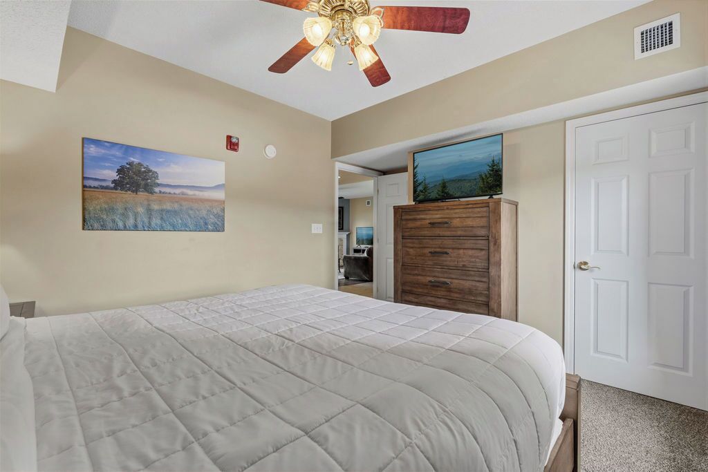 Une chambre à coucher avec un lit, un ventilateur de plafond, une commode en bois, une porte et un tableau accroché au mur.