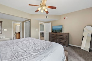 A bedroom with a bed, a dresser, a television, a ceiling fan, and a door leading to another room.