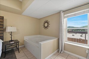 A room with a large bathtub, a small table with a lamp, a decorative mirror, and a window with a view of a parking lot. - Wake Up to the River, Walk to the Fun - 3 BR Condo (Pigeon Forge)