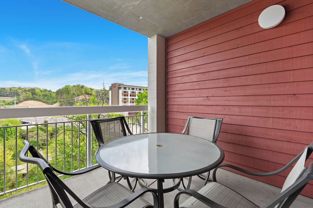 A balcony with a round table and four chairs, a red wall, and a view of greenery and buildings.