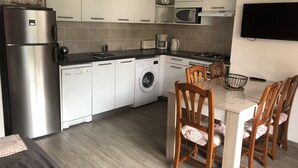 Eine kompakte Küche mit Kühlschrank, Mikrowelle, Spüle und Waschmaschine. Ein Esstisch mit Stühlen sowie ein Flachbildfernseher an der Wand sind vorhanden.