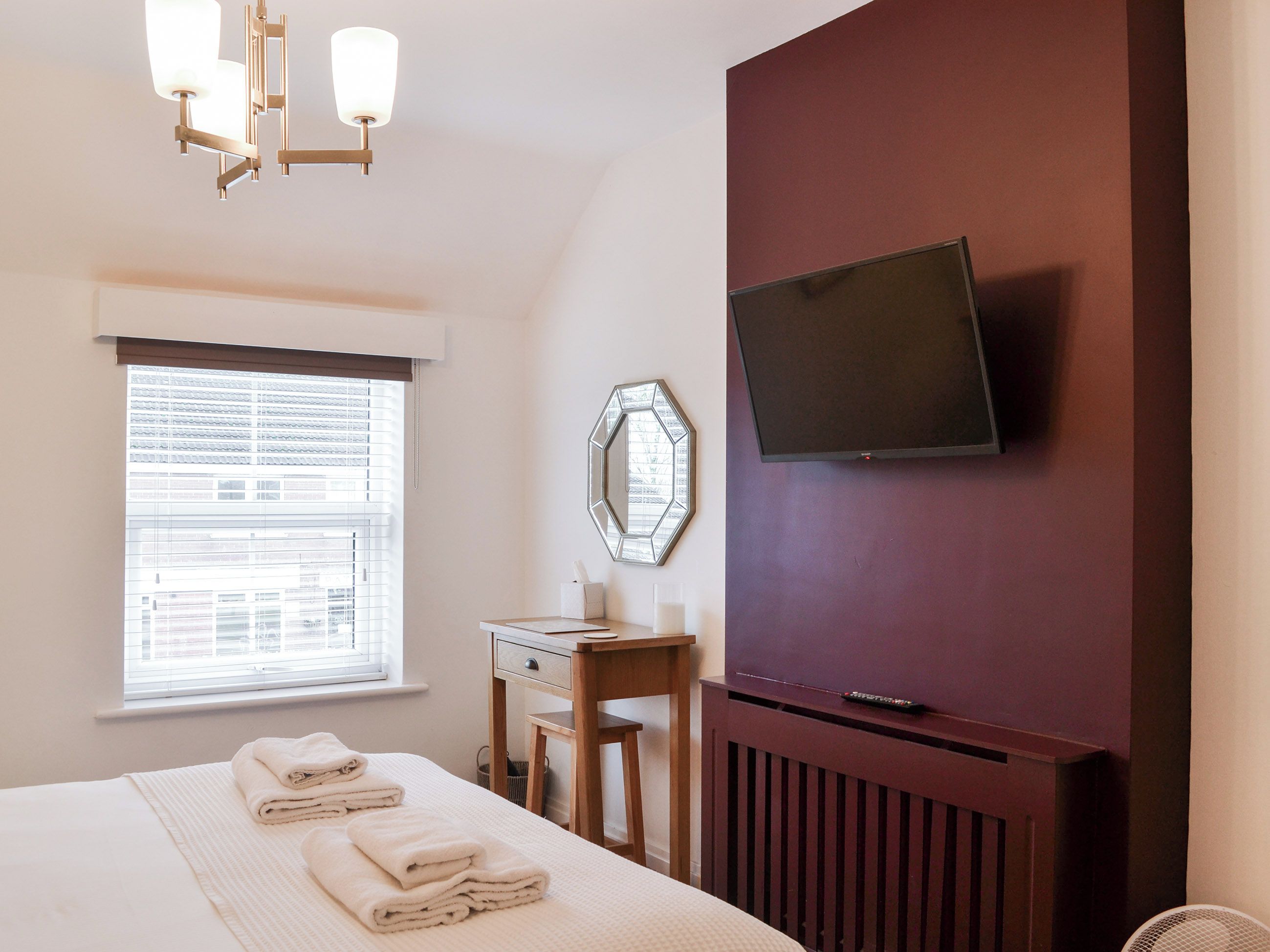 A hotel room with a bed, a television mounted on the wall, a window with blinds, and a bedside table with a mirror and a lamp.