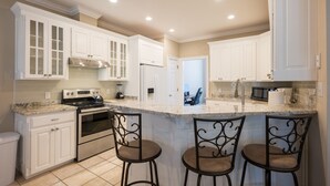 A kitchen with white cabinets, a stainless steel stove, and a marble countertop. There are three bar stools with metal frames.