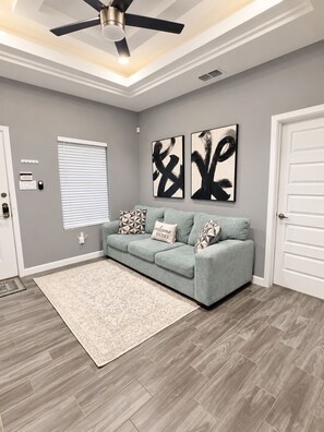 A modern living room with a grey sofa, abstract wall art, a ceiling fan, and a patterned rug.