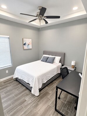 A bedroom with a bed, a desk, a chair, a ceiling fan, and a window with blinds.