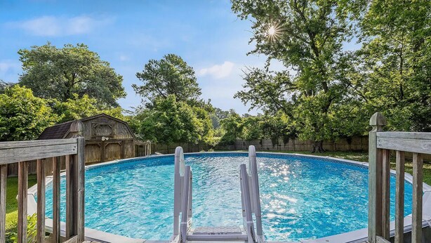 Una terraza de madera con una escalera de piscina que da a una piscina azul cristalina rodeada de árboles.