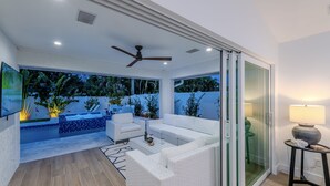 A modern living room with a white sofa, a coffee table, and a ceiling fan. There is a glass door leading to an outdoor area with a pool and plants. - Tee, Splash & Relax | Putting Green, Pool, Spa (Naples)