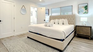 Een slaapkamer met een groot bed, een nachtkastje, een spiegel en een raam met jaloezieën.