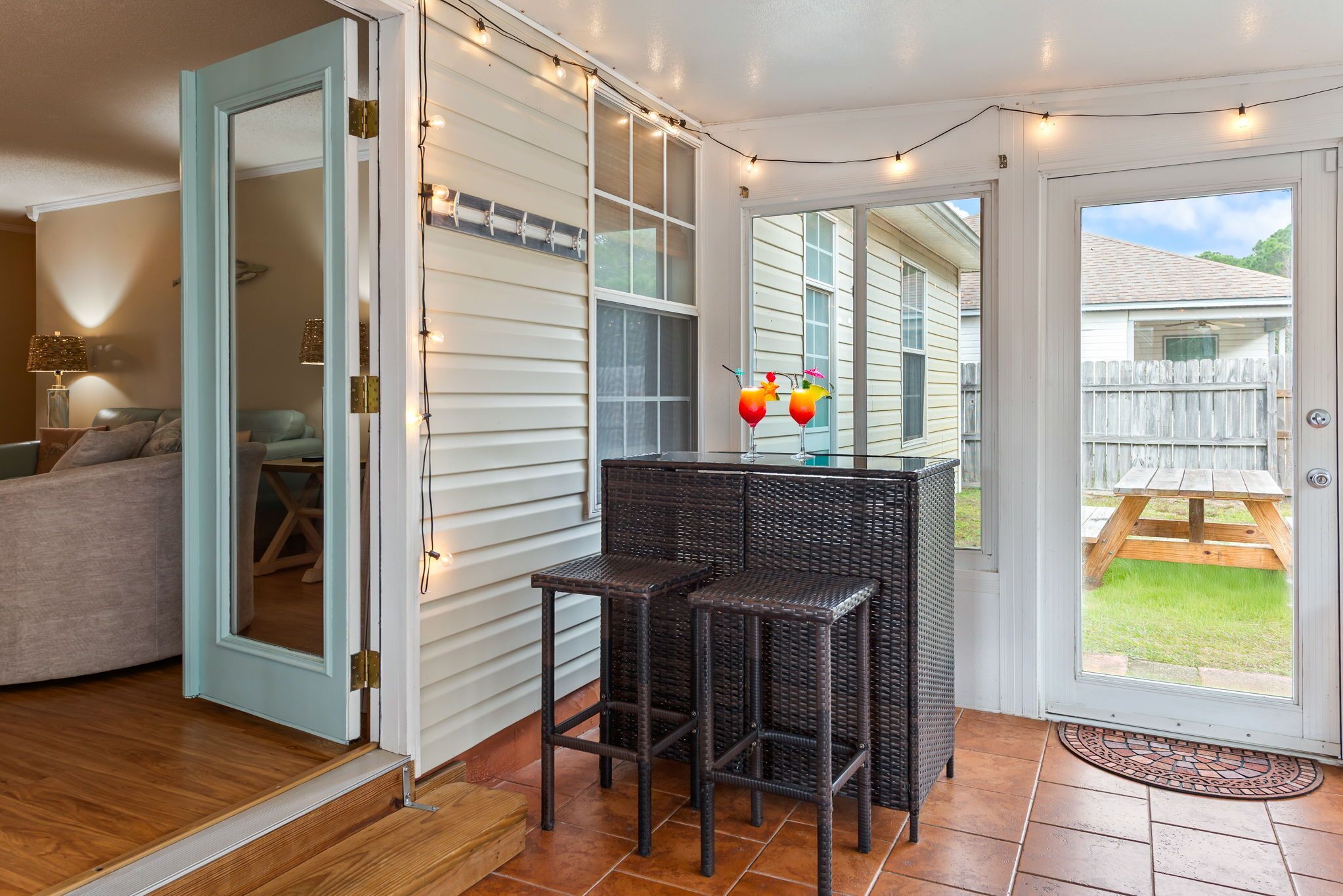 An open sliding glass door leading to a patio with a wooden table and chairs, string lights, and a wicker bar stool.