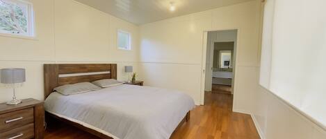Un dormitorio con cama de madera, mesitas de noche, una mesita auxiliar, una ventana y un pasillo que lleva a un baño.