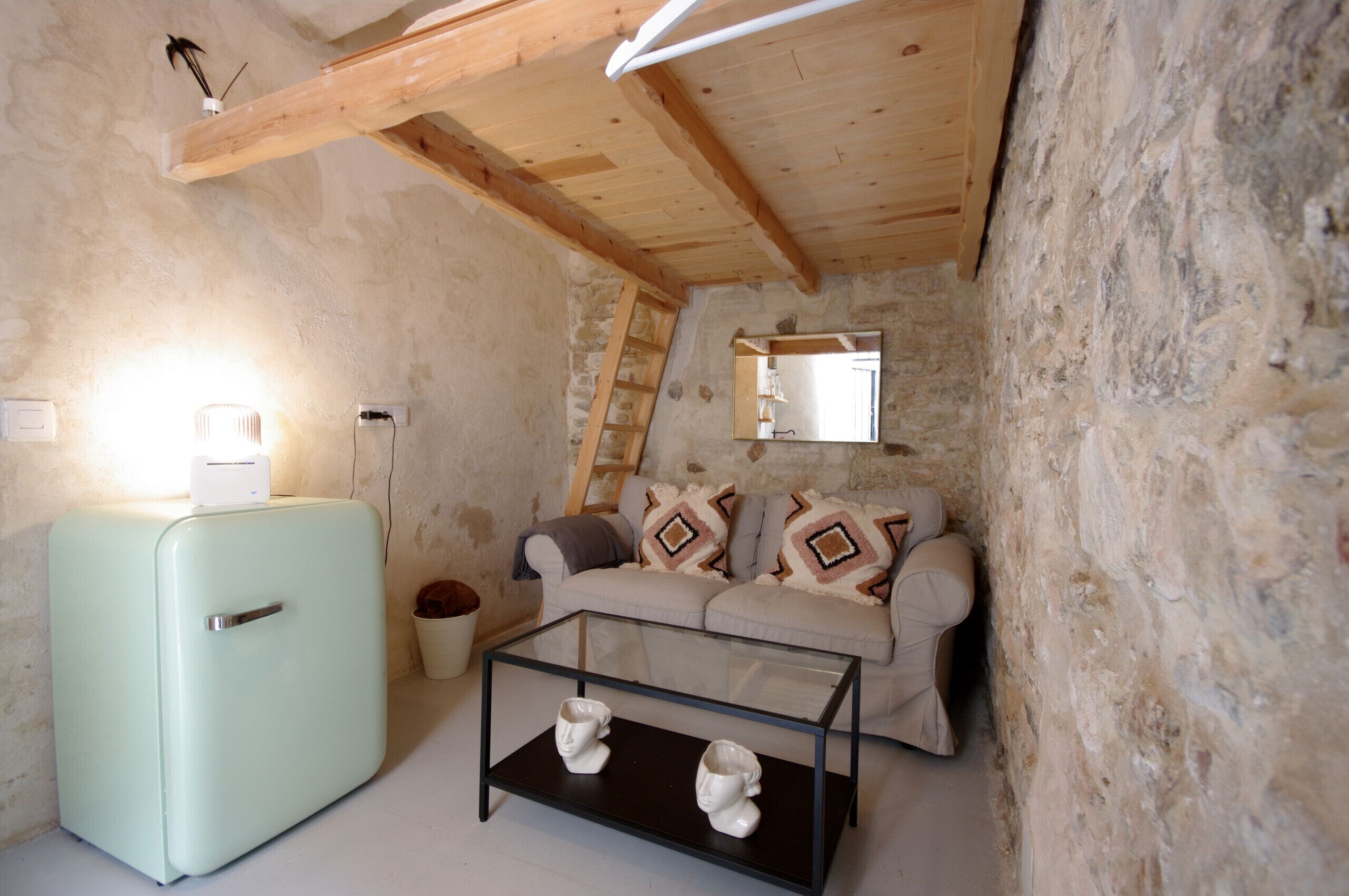A rustic room with a vintage light blue refrigerator, a glass-top coffee table with two white sculptures, a wooden sofa with patterned cushions, and a stone wall.