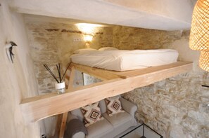 Una stanza con parete in pietra, struttura letto in legno, comodino con lampada e un piccolo angolo salotto con cuscini.