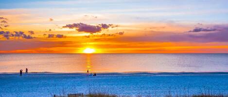 A beach with a sunset, calm water, and people walking.