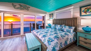 Une chambre à coucher avec un lit, un banc, une table de chevet, une lampe et une vue sur l’océan au coucher du soleil.