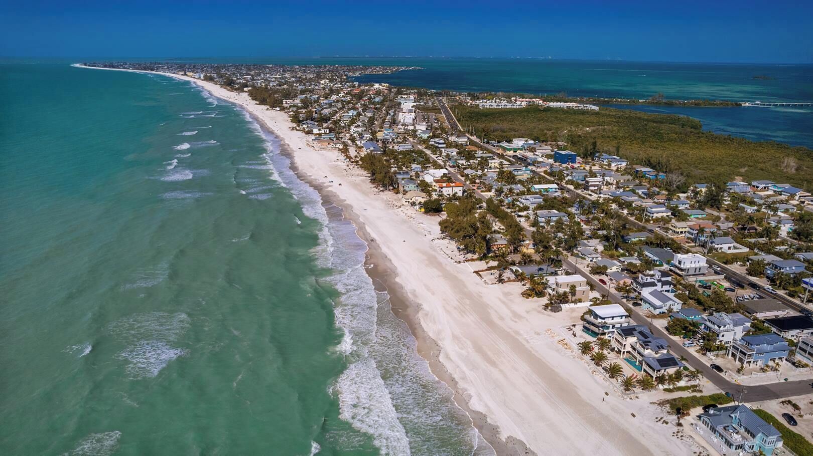 An aerial view of a coastal town with beachfront properties, roads, and a clear blue sky.
