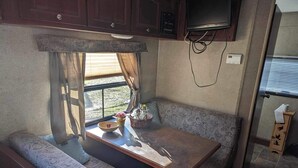 Une cuisine compacte pour camping-car, dotée d’une table et de sièges intégrés, d’un téléviseur fixé au-dessus du comptoir et d’une fenêtre laissant entrer la lumière naturelle.
