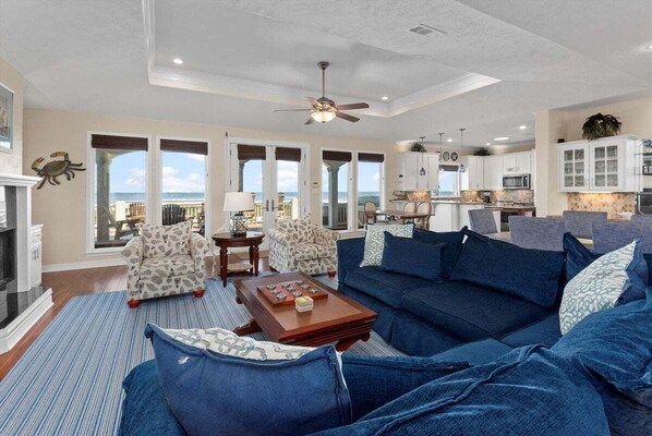 A spacious living room with a large blue sofa, a patterned armchair, and a coffee table. There is a view of the ocean through large windows.
